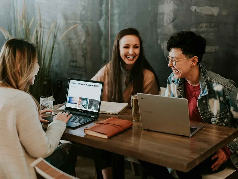 Três estudantes conversando e rindo em volta de uma mesa com notebooks